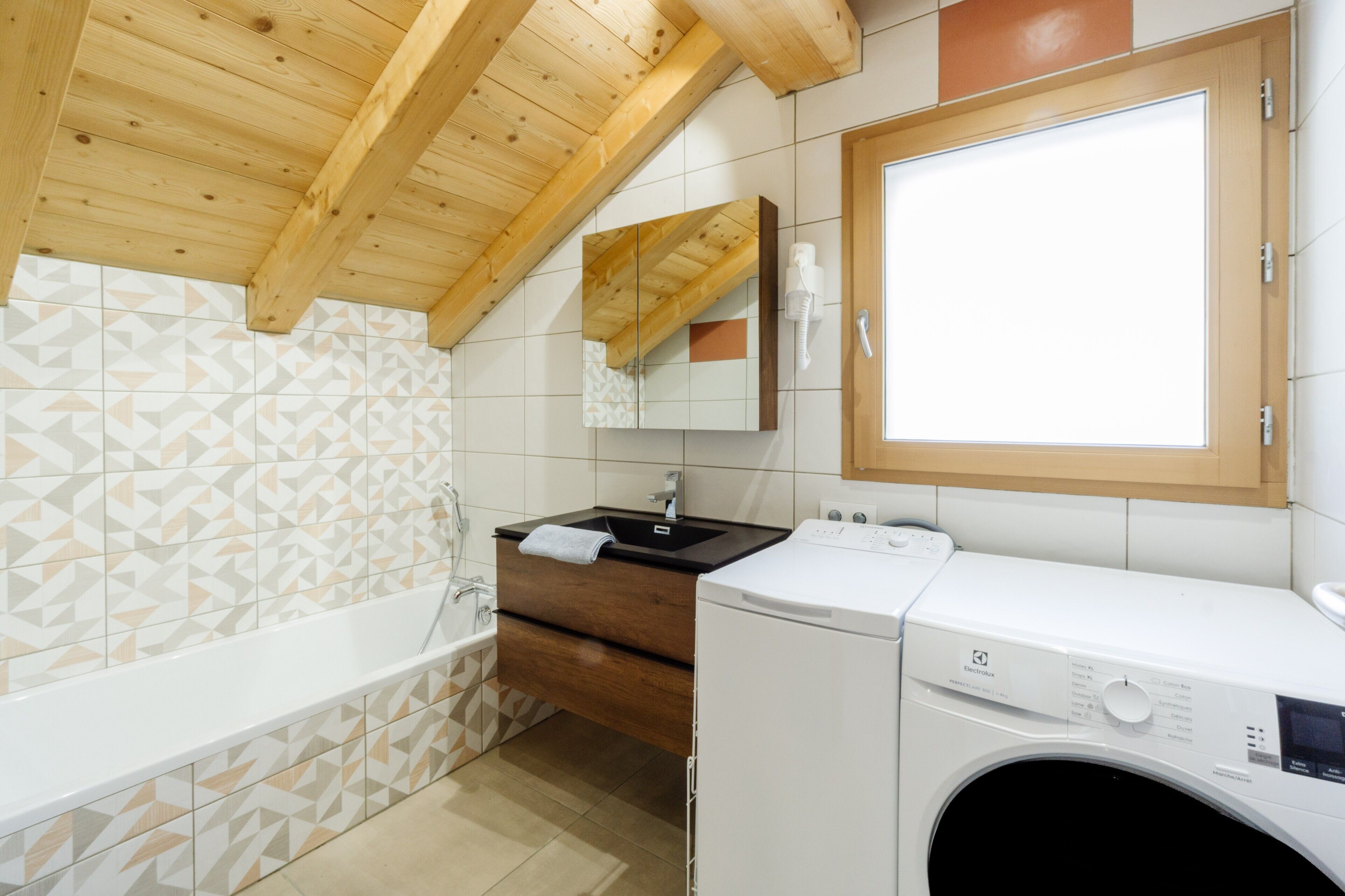 A modern bathroom with a wooden ceiling, a bathtub, a sink with a mirror, and a washing machine.