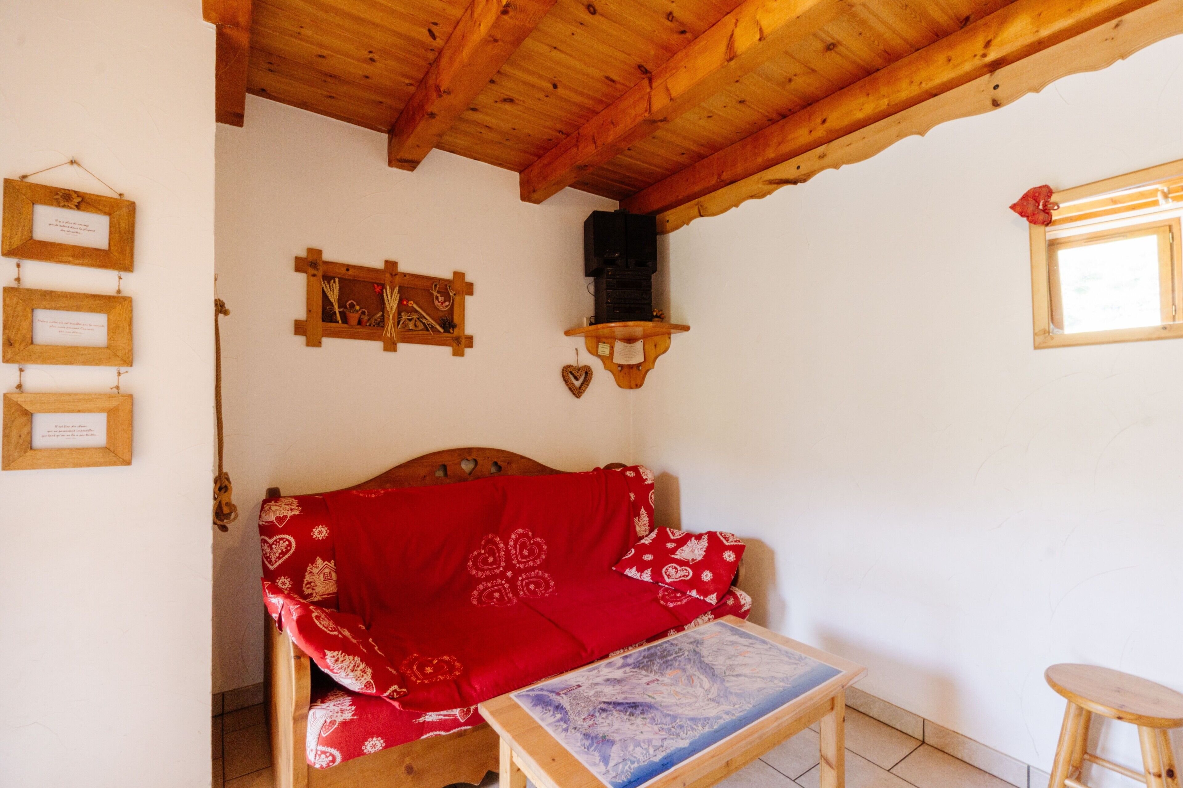 Una habitación acogedora con techo de madera, una cama con manta roja, una mesita con mantel azul y un estante de madera con un altavoz.