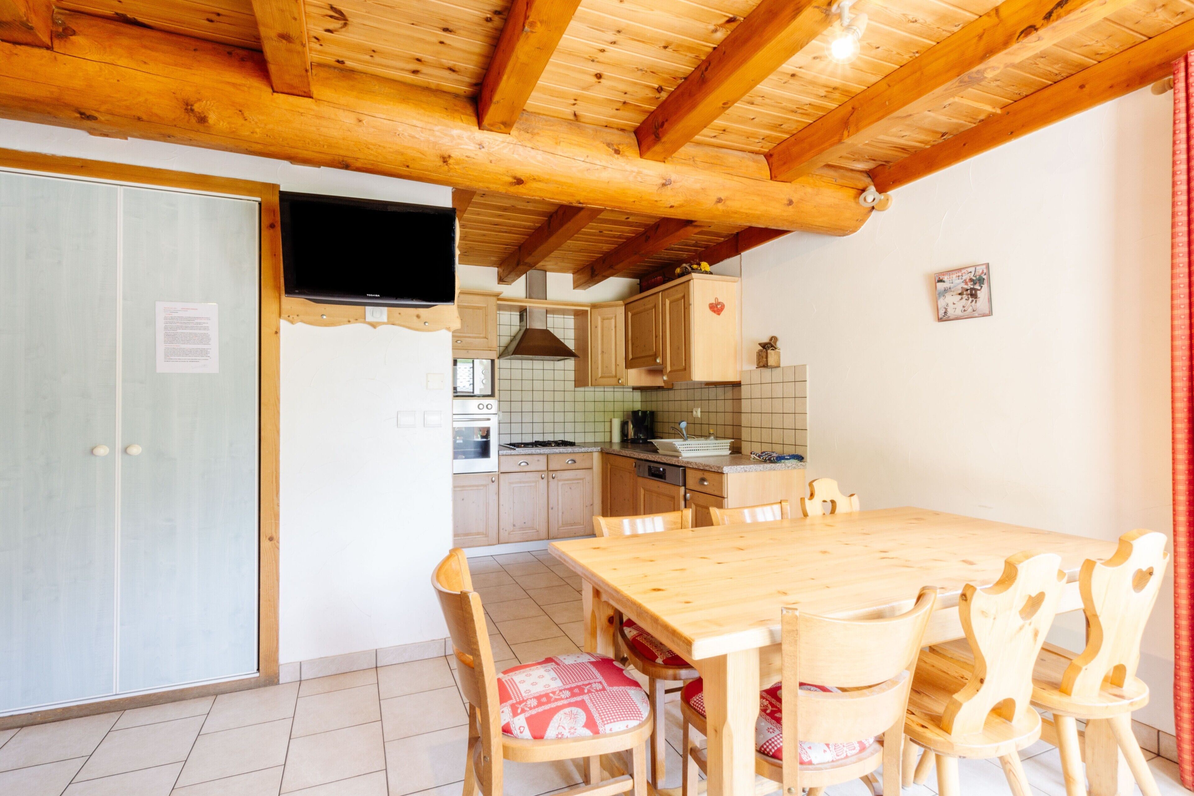 Ein Holztisch mit Stühlen, eine Küchenzeile mit Schränken und einem Herd sowie ein an der Wand befestigter Fernseher.