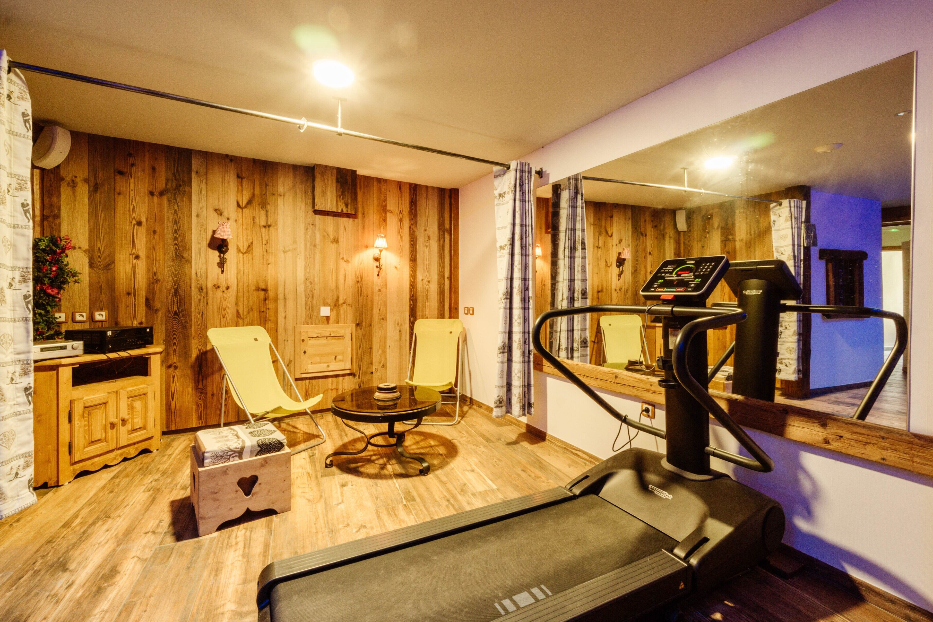 Une pièce équipée d’un tapis de course, avec des murs en bois, un miroir et une petite table accompagnée d’une chaise.