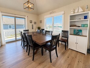 A dining area with a wooden table and chairs, a chandelier, and a view through the windows. - SEA GLASS SHACK -  4 bed, 2 bath, Pool, Hot Tub & Dogs Allowed (Corolla)