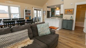 A cozy living room with a sofa, a dining area with chairs, a kitchen with a refrigerator, and a bookshelf. - SEA GLASS SHACK -  4 bed, 2 bath, Pool, Hot Tub & Dogs Allowed (Corolla)