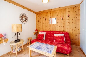 Una habitación acogedora con pared de madera, un sofá rojo con estampado, una mesita redonda con lámpara y un cuadro en la mesa.