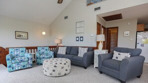 Una sala de estar con un sofá gris, un sillón azul estampado y un banquito redondo. Hay una lámpara sobre una mesita auxiliar y un cuadro en la pared.