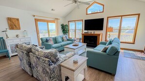 A spacious living room with a large TV, a fireplace, and a balcony with a view. - Dunes Crest - Ocean and sound views in Duck, NC! 5 bed, 4.2 bath, pool, hot tub (Duck)