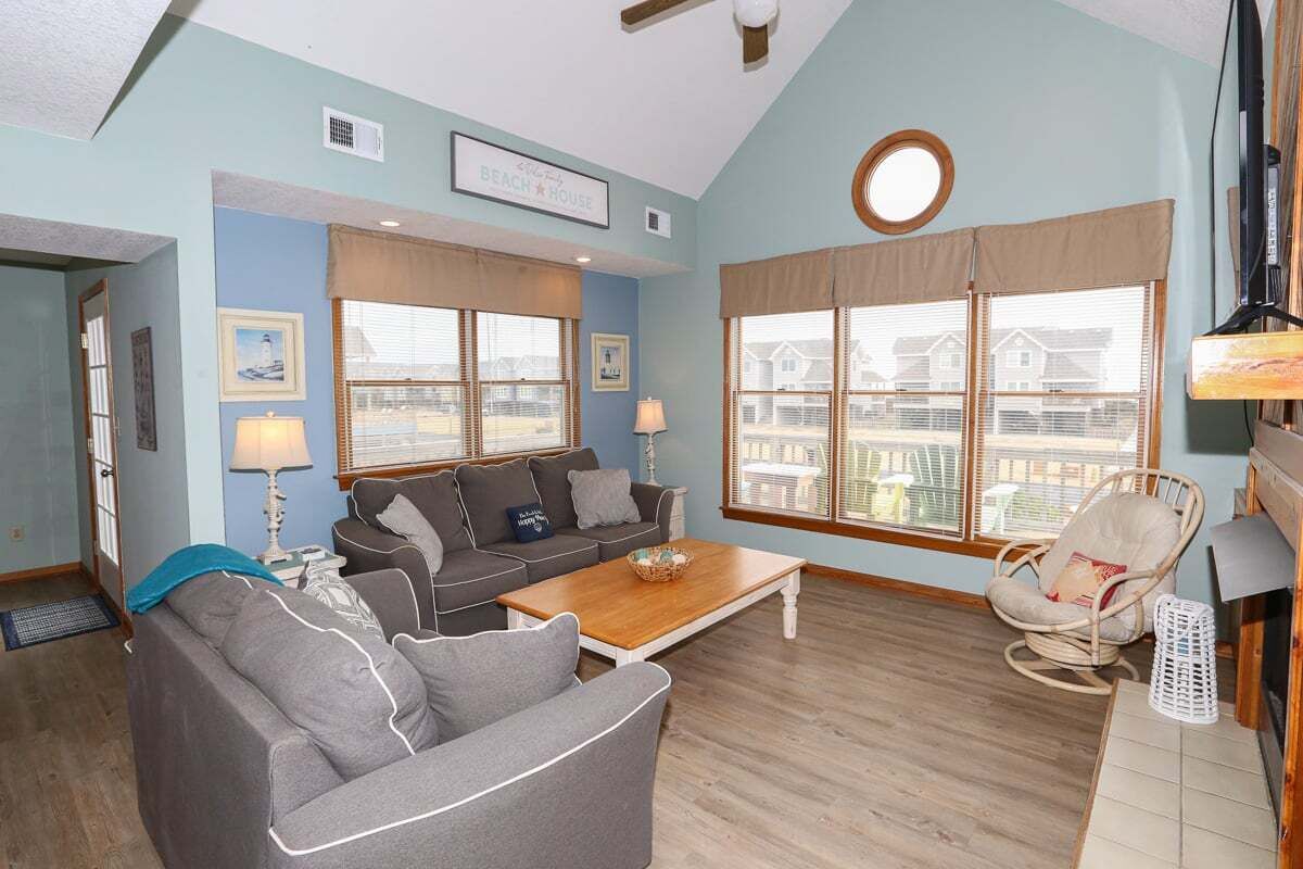 A living room with a sofa, a coffee table, and a chair. There are two windows with blinds, a ceiling fan, and a sign on the wall.