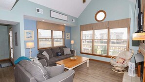 A living room with a sofa, a coffee table, and a chair. There are two windows with blinds, a ceiling fan, and a sign on the wall. - PELICAN'S LANDING - Beautiful semi-oceanfront townhouse with community pool, Great location! (Southern Shores)