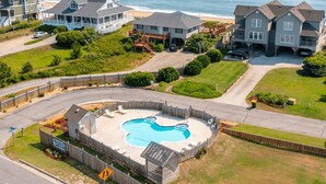 A residential area with a swimming pool surrounded by a wooden fence, houses in the background, and a road in the foreground. - PELICAN'S LANDING - Beautiful semi-oceanfront townhouse with community pool, Great location! (Southern Shores)