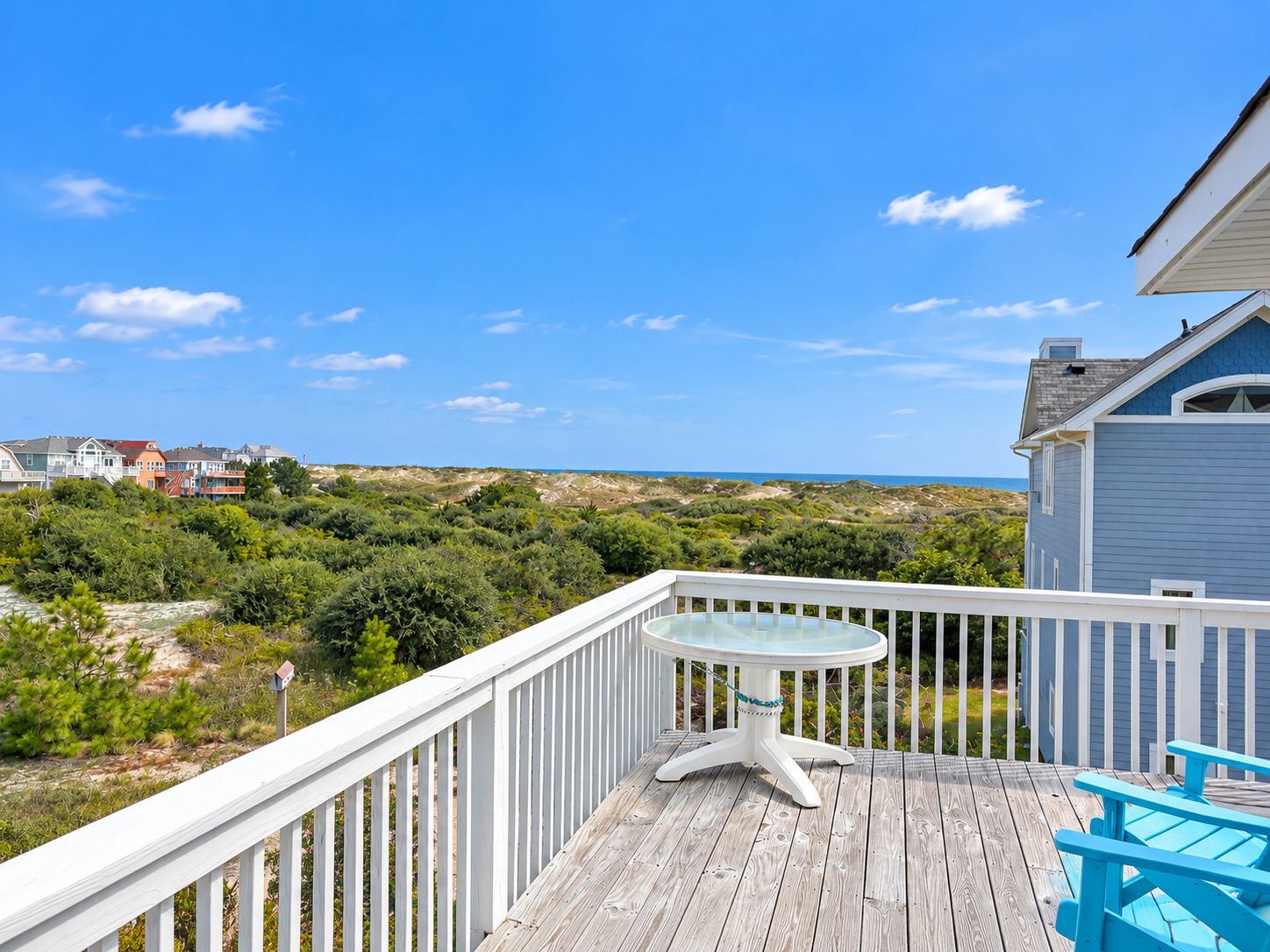 A wooden deck with a glass table and chairs, overlooking a coastal landscape with houses and vegetation.
