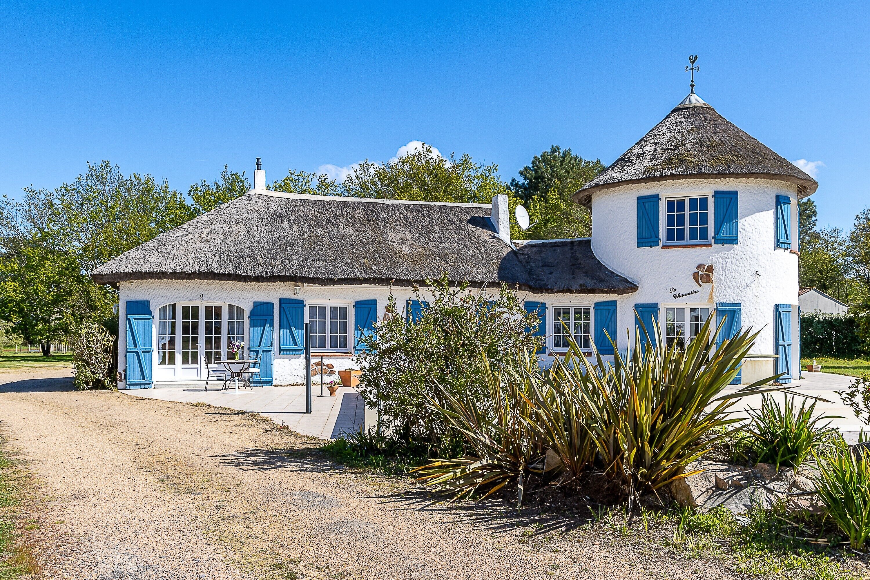 Un bâtiment blanc avec des volets bleus et un toit de chaume, entouré de verdure.
