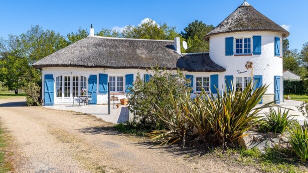 Une maison à deux étages, avec un toit de chaume, des murs blancs et des volets bleus.