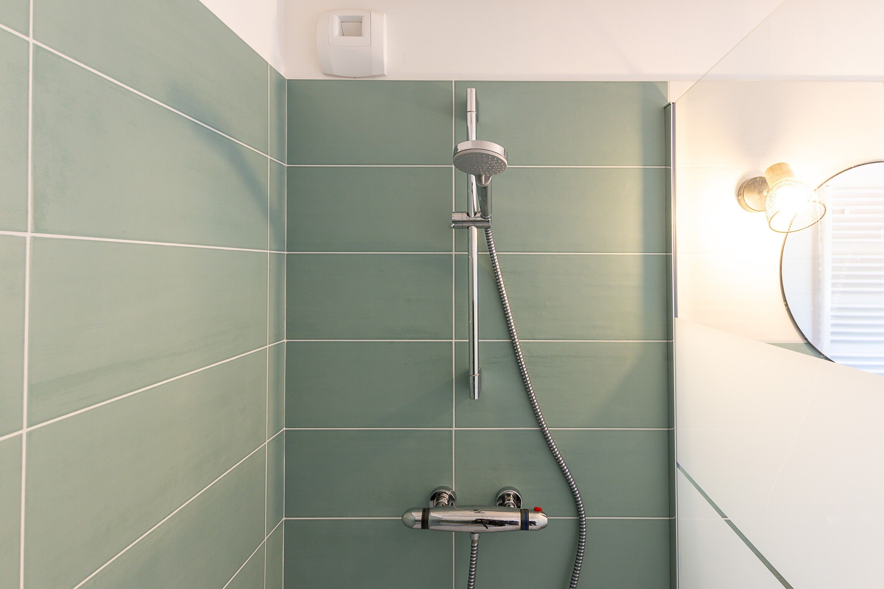 A shower with a handheld showerhead and a wall-mounted faucet, green tiled walls, and a light fixture on the wall.