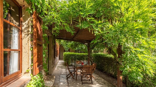 A covered outdoor seating area with stone flooring, wooden furniture, and lush greenery. - Apartment 'Il Covone' with Shared Pool, Shared Terrace and Wi-Fi (Rapolano Terme)
