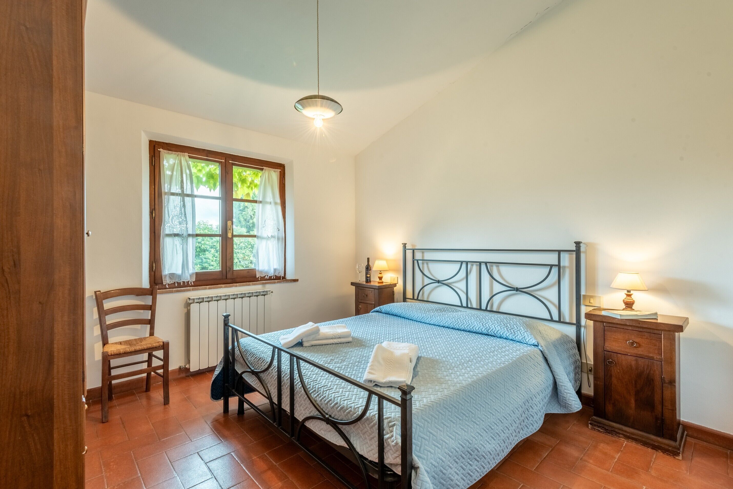 A bedroom with a bed, a chair, a nightstand with a lamp, a window with curtains, and a radiator.