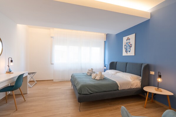 Une chambre à coucher moderne, avec un lit, un bureau et une chaise, une fenêtre avec des rideaux et une œuvre d’art accrochée au mur.