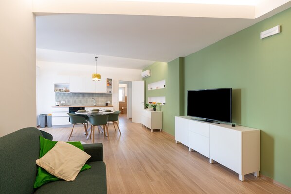 A modern living room with a green sofa, a flat-screen TV, and a dining area with a table and chairs.