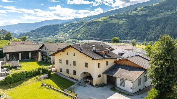 A large, two-story building with a sloped roof, surrounded by lush greenery and mountains in the background.