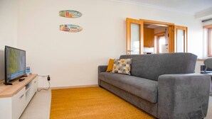 A living room with a grey sofa, a television, a small table, and a yellow rug. - Apartment 'Borealis  - Nazaré' with Sea View, Shared Terrace and Wi-Fi (Nazaré)