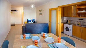 A dining area with a table set for four, a washing machine, and a television in the background. - Apartment 'Borealis  - Nazaré' with Sea View, Shared Terrace and Wi-Fi (Nazaré)