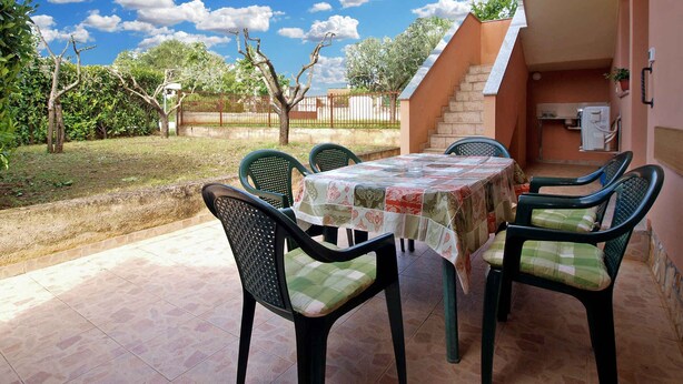 An outdoor patio with a table and chairs, a staircase leading to an upper level, and a view of a fenced garden with trees and a clear sky.
