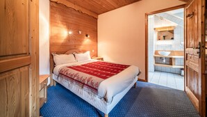 Quarto com teto de madeira, cama com cobertor vermelho e branco, mesa de cabeceira e guarda-roupa de madeira.
