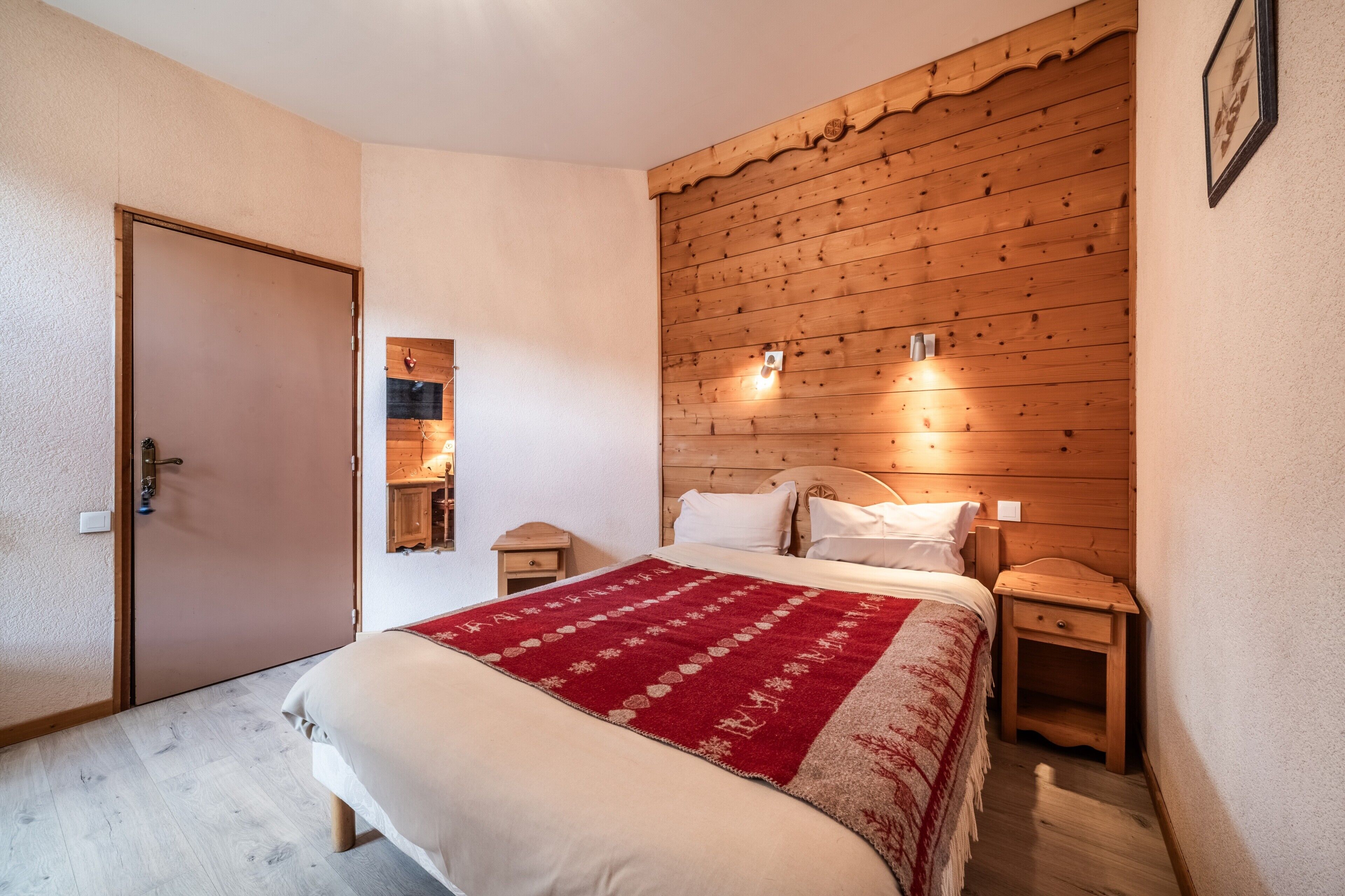 A bedroom with a wooden headboard, a bed with a red and white blanket, bedside tables, and a door.