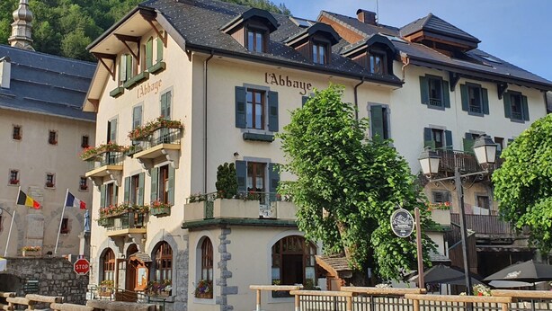 A building with the sign 'l'Abbaye' indicating it is a hotel. It features a balcony with flowers, a stone facade, and a mountain backdrop.
