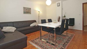 Een moderne woonkamer met een donkerbruine bank, een witte eettafel met vier stoelen, een kamerkleed met een patroon en een hanglamp.