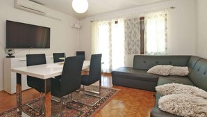 Een moderne woonkamer met een eethoek, een flatscreen televisie aan de muur, een grijze bank met zachte kussens en een vloerkleed met een patroon op de houten vloer.