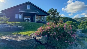 Une maison moderne dotée d’un grand porche, entourée d’un jardin où poussent des fleurs roses et où l’on trouve de gros rochers.