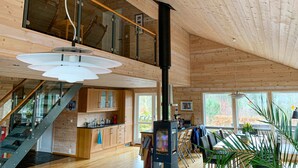 Un intérieur spacieux en bois, doté d’une cheminée centrale, d’une mezzanine et de grandes fenêtres offrant une vue sur l’extérieur.