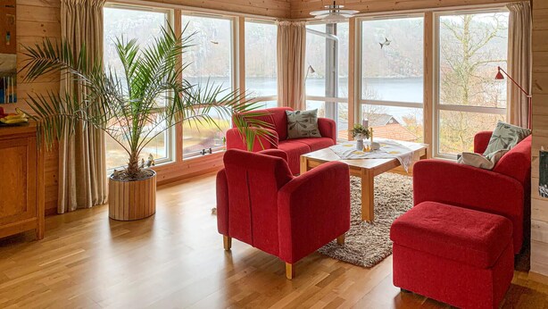 Une pièce chaleureuse avec un canapé rouge, un plancher en bois et de grandes fenêtres donnant sur une étendue d’eau.