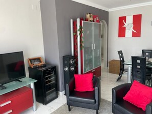 Un salon comprenant un téléviseur, un bureau avec un plateau en verre, un meuble rouge, un fauteuil en cuir noir et un coin repas avec une table et des chaises.