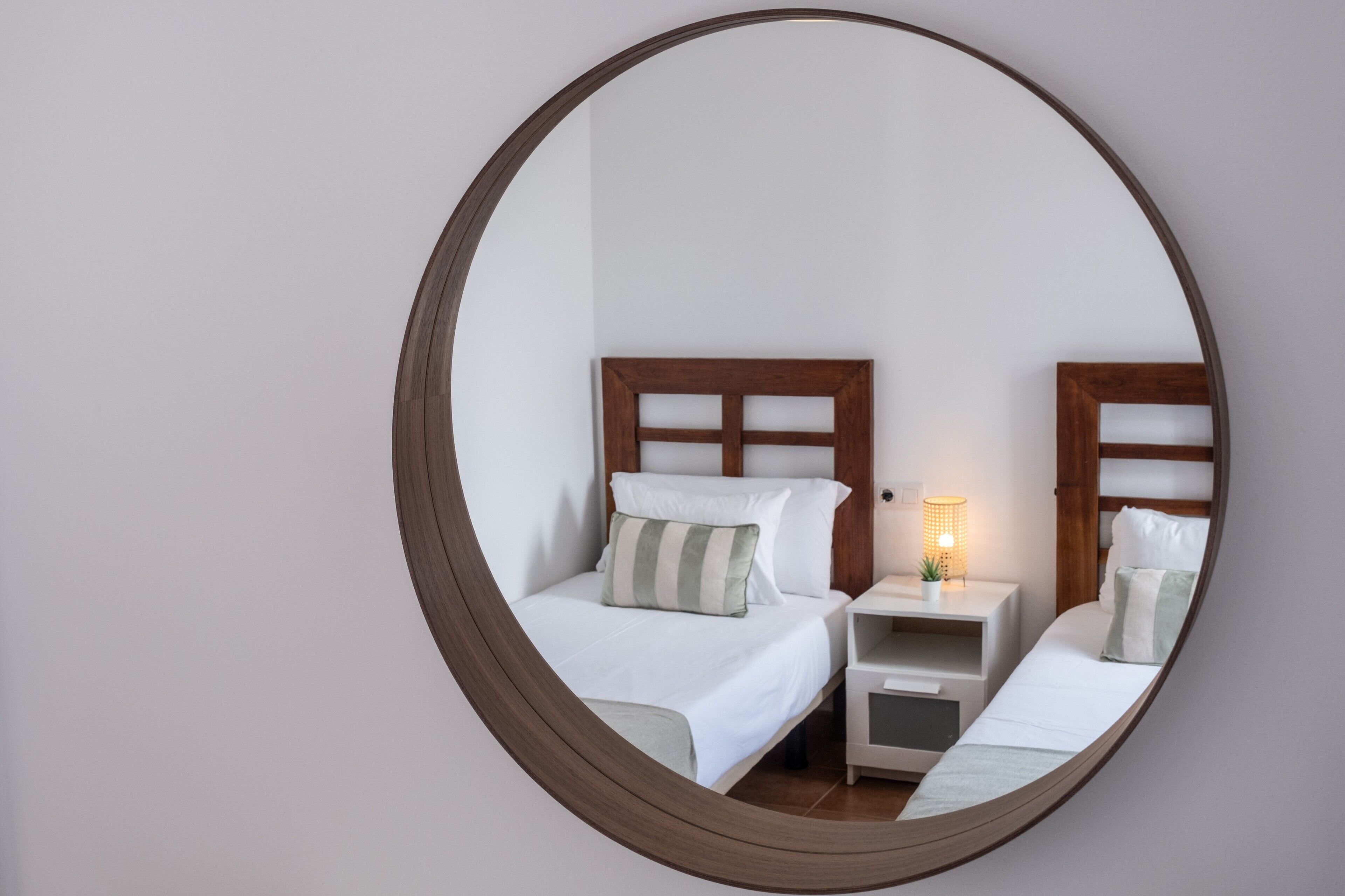 A circular mirror reflecting a bedroom with two beds, a bedside table with a lamp, and a small plant.