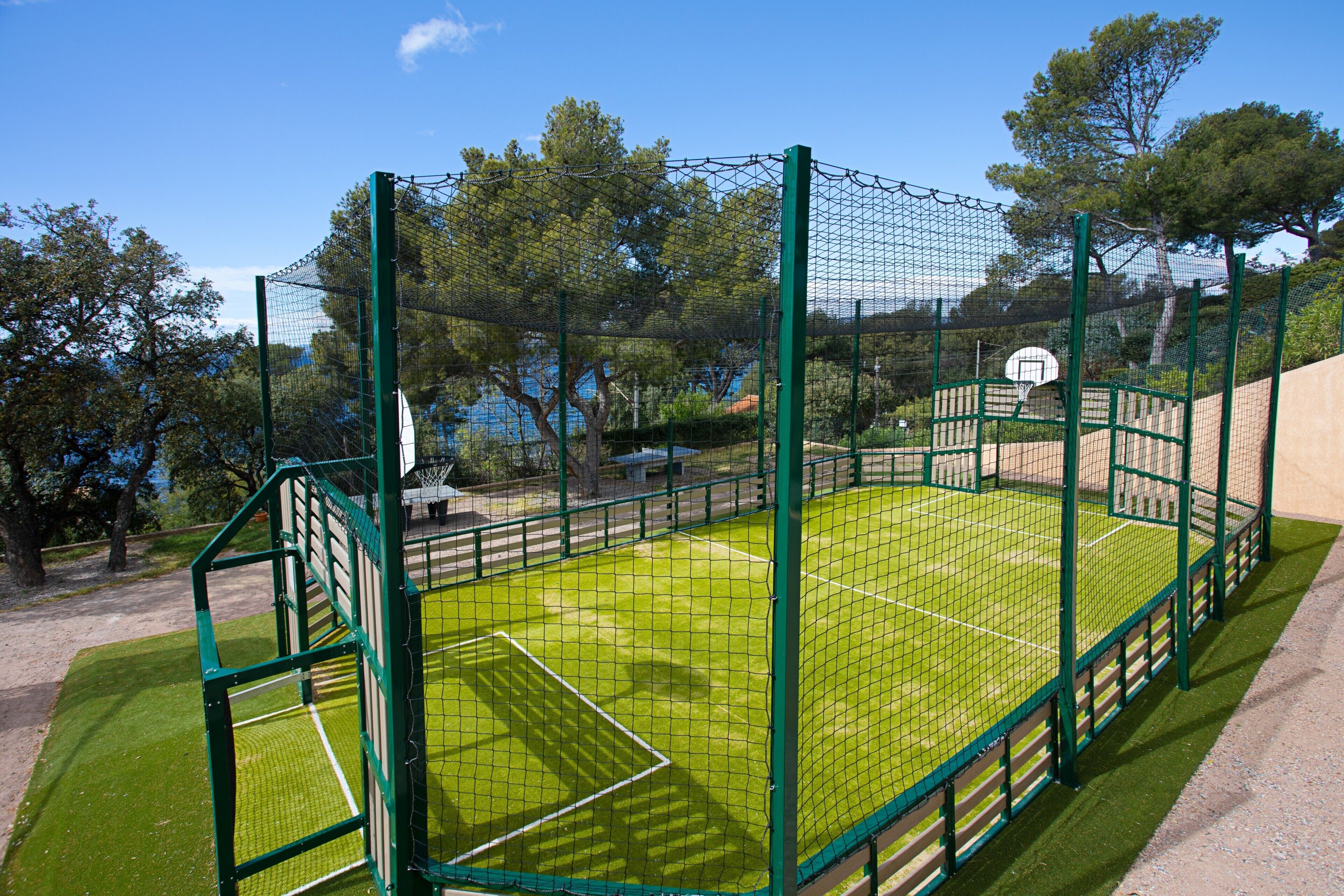 Een sportveld met een basketbalring, omgeven door een groene hekken en bomen op de achtergrond.