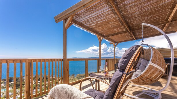 A wooden deck with a hanging chair, a table with a glass, and a view of the sea.