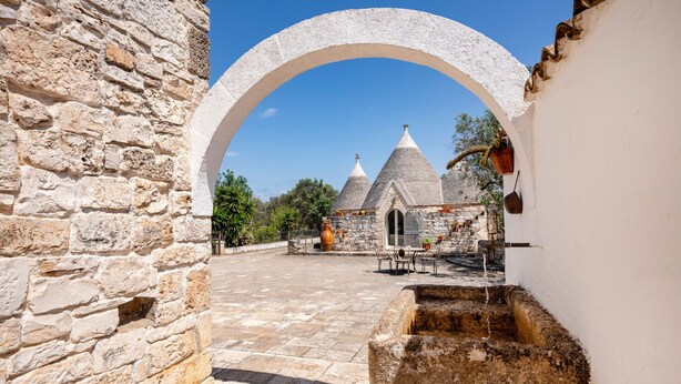 Un arc en pierre mène à une cour intérieure agrémentée d’une fontaine, de murs en pierre et d’un bâtiment surmonté de toits coniques.