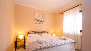 Una cama bien hecha con edredón blanco y dos almohadas, una mesita de noche con lámpara, un cuadro en la pared y una ventana con cortinas traslúcidas.