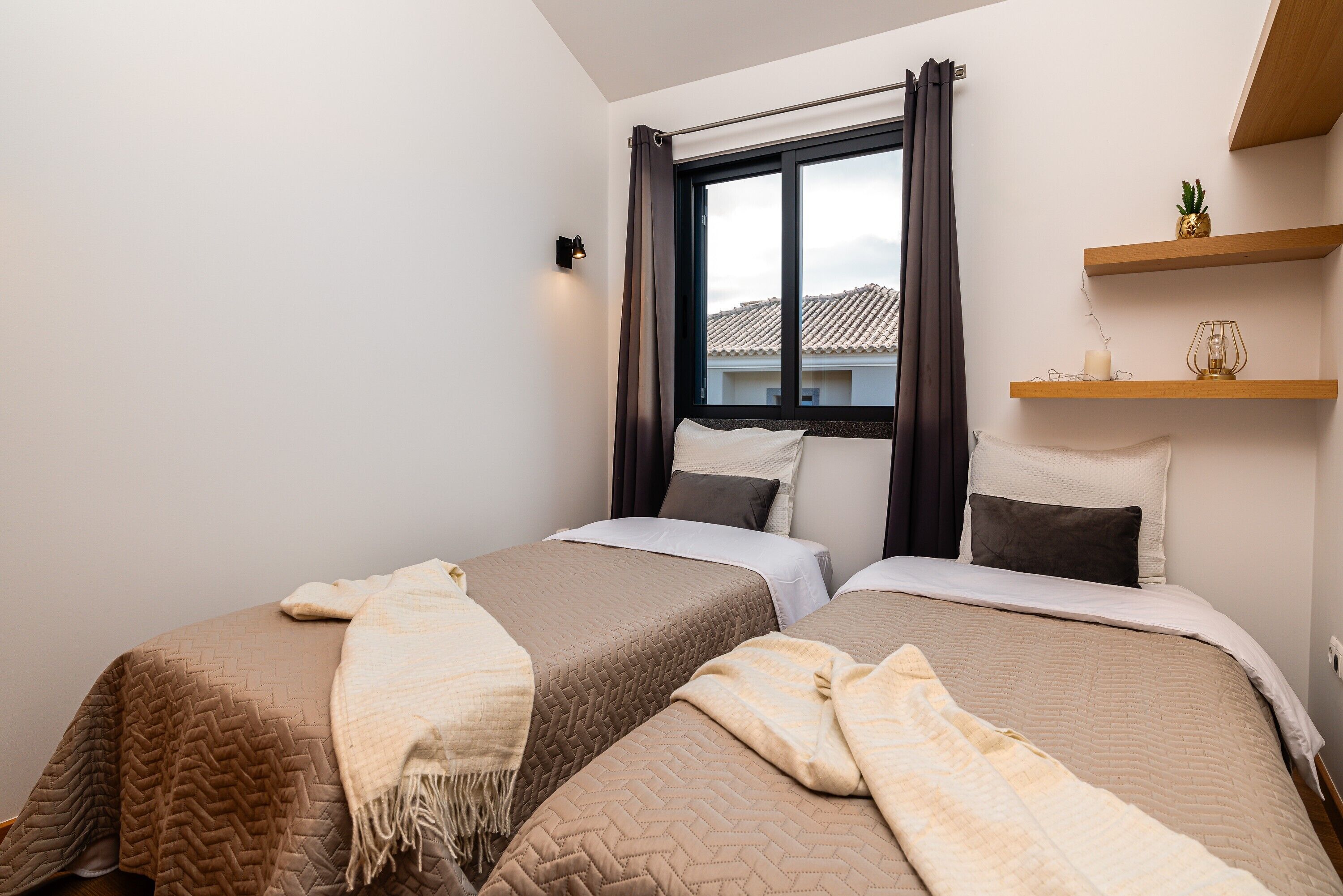 Une chambre avec deux lits, une fenêtre avec des rideaux, une étagère avec des objets décoratifs et un luminaire fixé au mur.