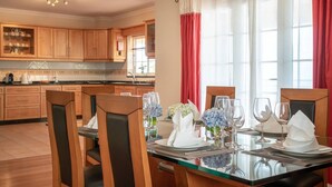A dining area with a glass table set for six, wooden chairs, and a view of the kitchen in the background.