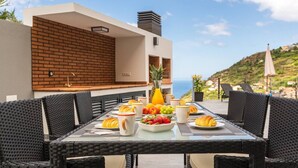 A dining table with breakfast items, wicker chairs, a modern kitchen with a brick wall, and a view of the sea.