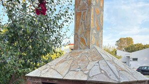 A stone-built outdoor pizza oven with a chimney, surrounded by a garden with trees and a clear sky.