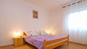 A bedroom with a wooden bed, two bedside tables with lamps, a purple bedspread, a wooden floor, and a window with curtains.