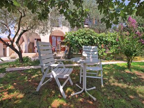 Un jardin avec deux chaises blanches et une table, entouré de verdure et, au fond, un bâtiment orné d’arcades.