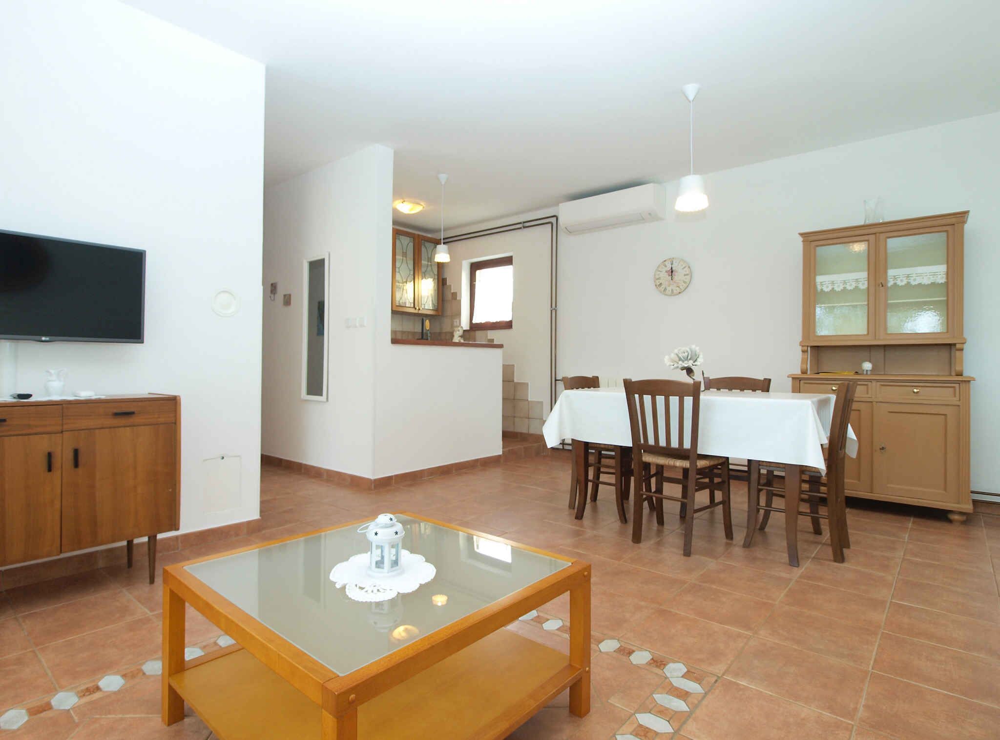 A spacious living area with a glass coffee table, wooden dining table, and chairs. A television is mounted on the wall, and there is a cabinet with glass doors.