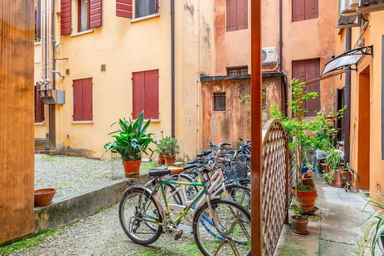 A narrow alley with bicycles parked along the side, potted plants, and buildings with red shutters.