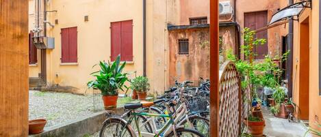 A narrow alley with bicycles parked along the side, potted plants, and buildings with red shutters.