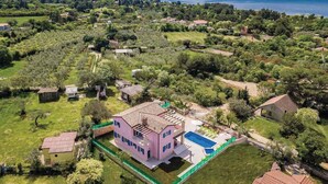 An aerial view of a residential area with a large pink house featuring a swimming pool, surrounded by greenery and a lake in the background. - Villa with swimming pool, terrace and barbecue (Peroj)