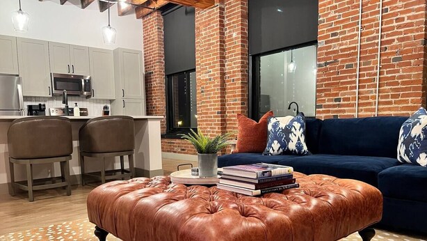 A modern kitchen with a brick wall, a dining area with two chairs, and a living room with a blue sofa and patterned cushions.