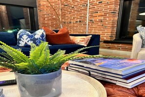 A living room with a brick wall, a blue sofa with patterned cushions, a potted fern, and a stack of books. - Brick & Bed Loft - City Views - Convention Center (St. Louis)